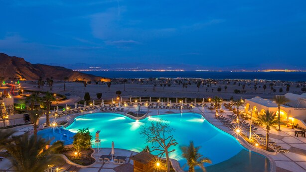 Una piscina di un resort con lettini e palme, circondata da un paesaggio desertico e vista sulla spiaggia di notte.