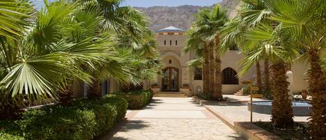A pathway leading to a building with arched doorways, surrounded by palm trees and a mountainous backdrop.