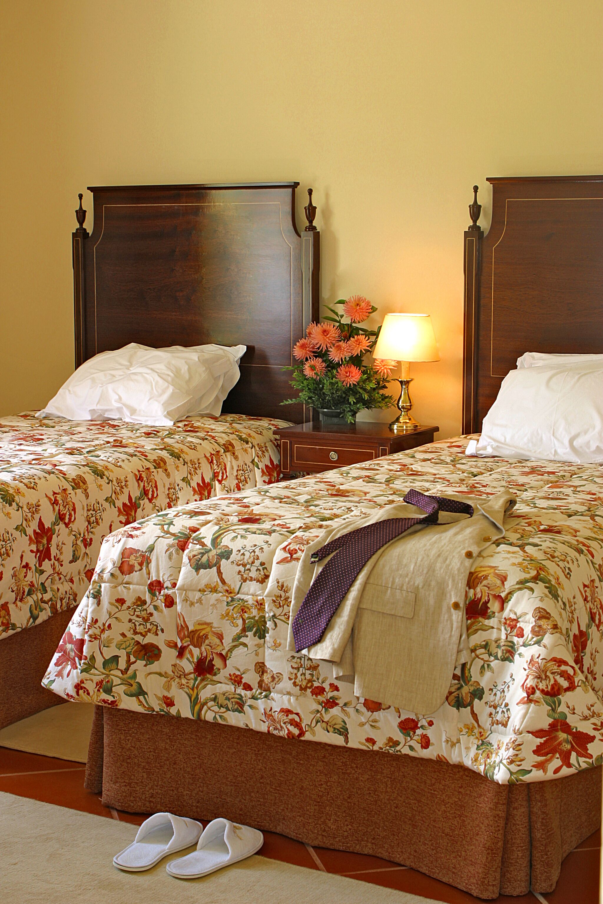 A hotel room with two beds, floral bedding, a nightstand with a lamp, and a pair of slippers on the floor.
