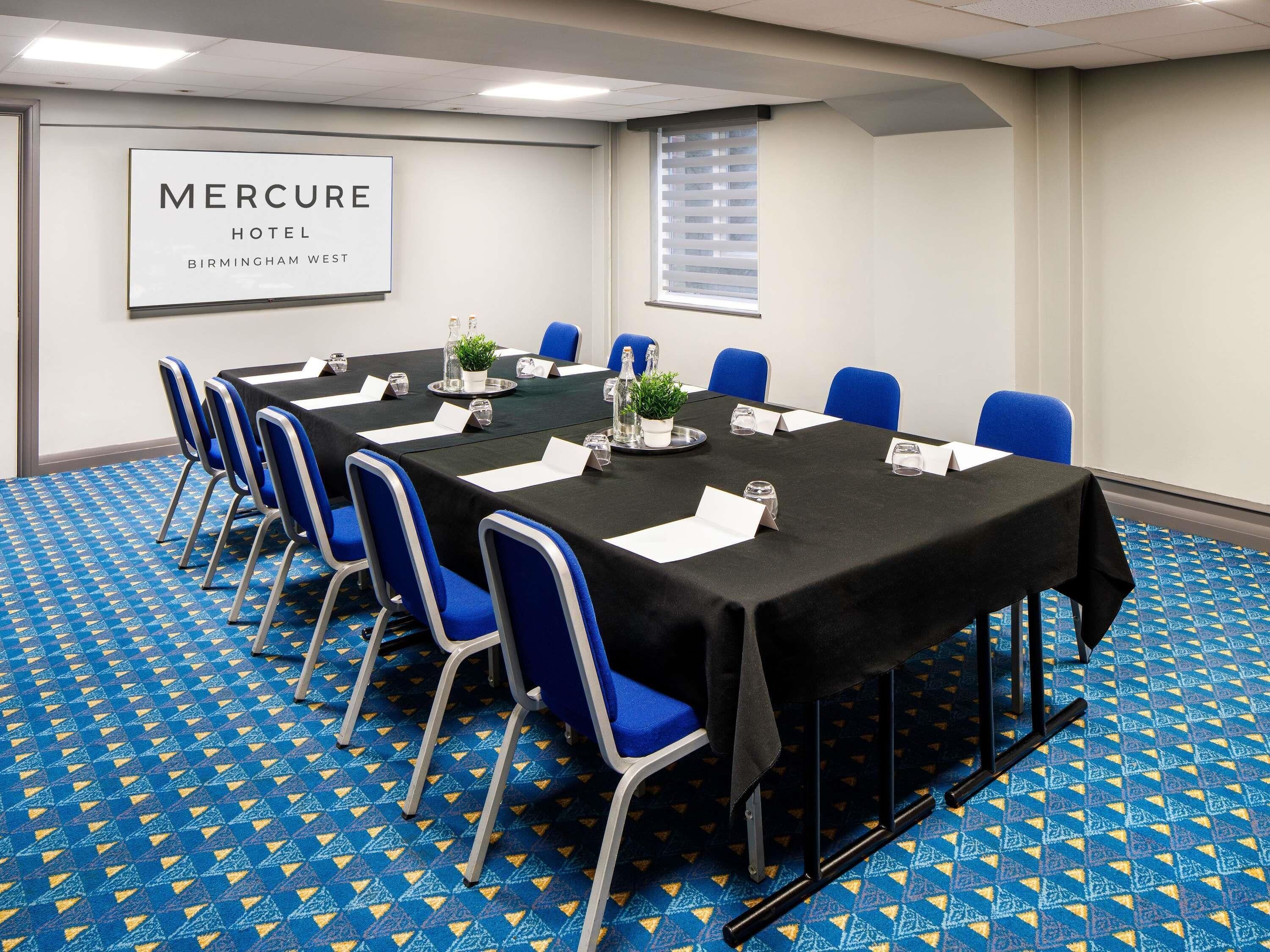Een vergaderruimte met een lange tafel, blauwe stoelen, plaatskaarten en een bord met de tekst Mercure Hotel Birmingham West.