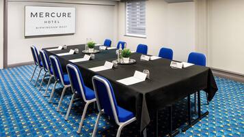 Een vergaderruimte met een lange tafel, blauwe stoelen, plaatskaarten en een bord met de tekst Mercure Hotel Birmingham West.
