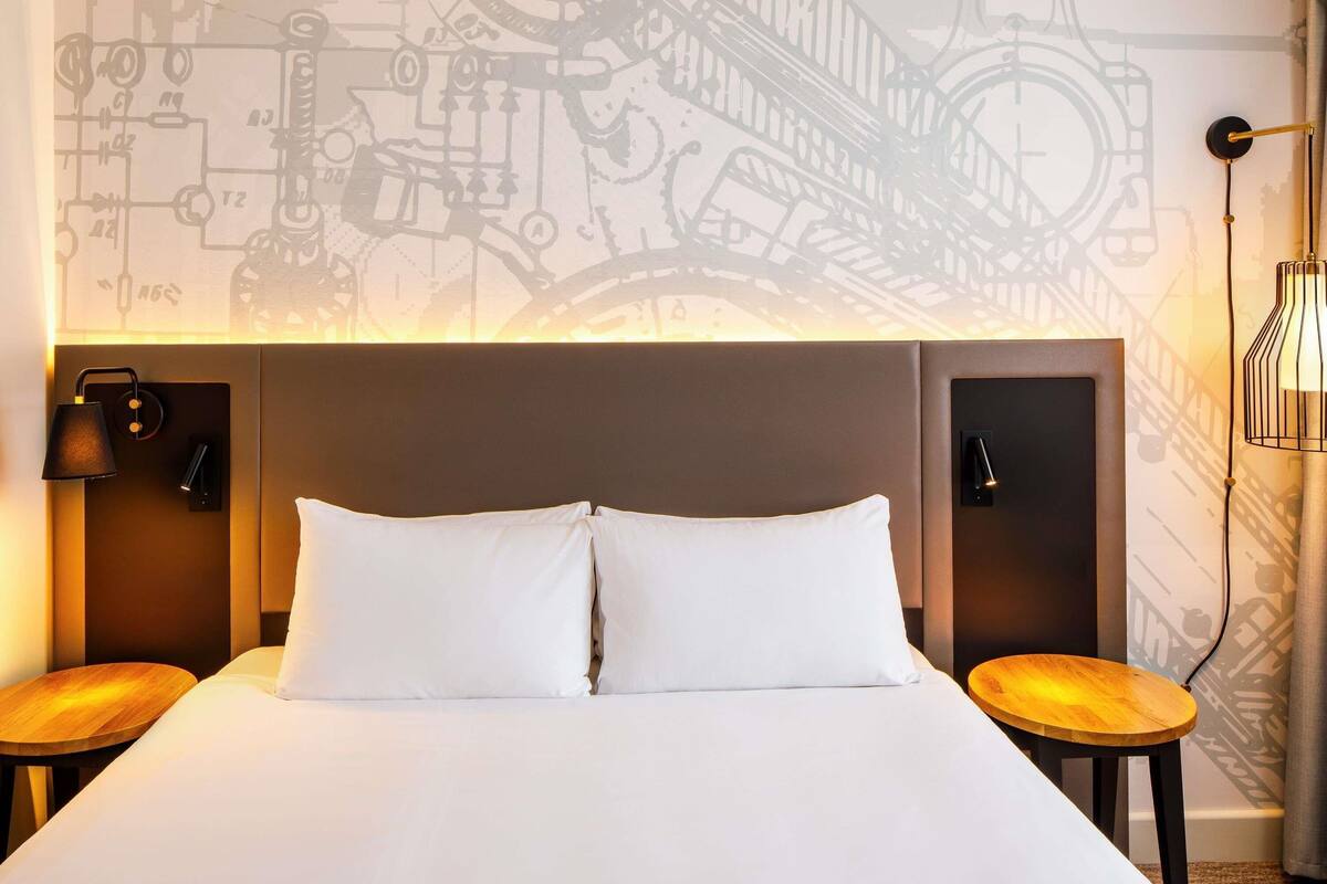 Habitación de hotel con una cama grande, dos mesitas de noche, cabecero con lámpara y pared con un mural de diseño moderno.