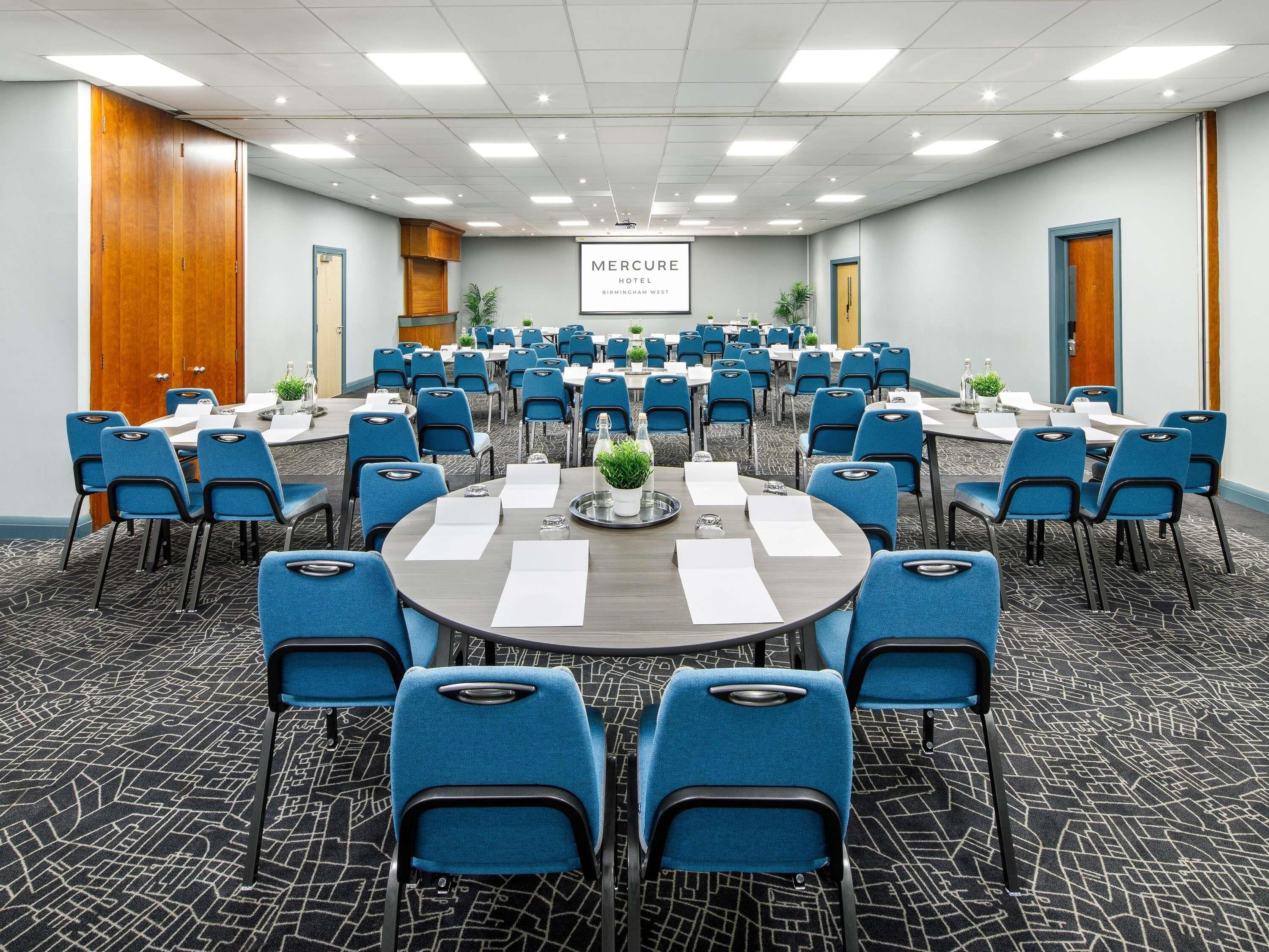 Un salón de conferencias con mesas redondas, sillas azules y un cartel que indica 'Mercure Hotel Birmingham West'.