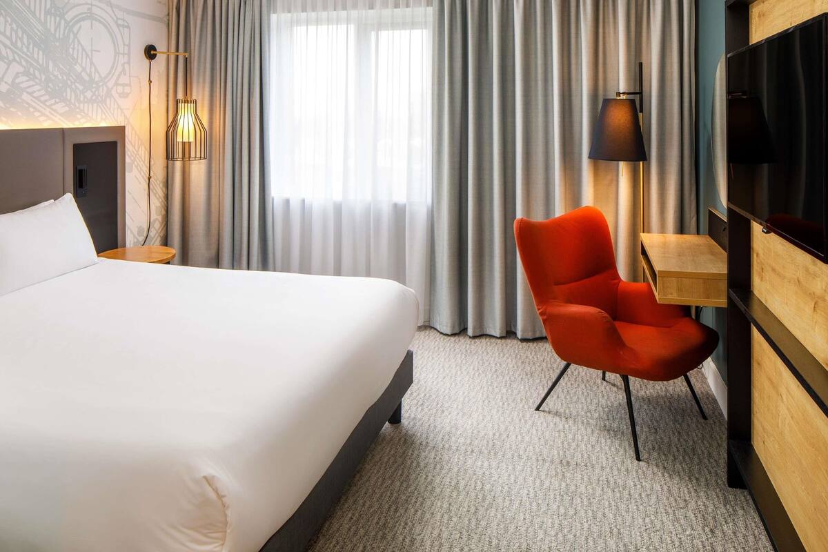 Habitación de hotel con una cama grande, una silla roja, un escritorio, un televisor y una ventana con cortinas.