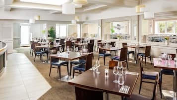 En rummelig restaurant med flere spiseborde og stole, store vinduer og hængende pendellamper.