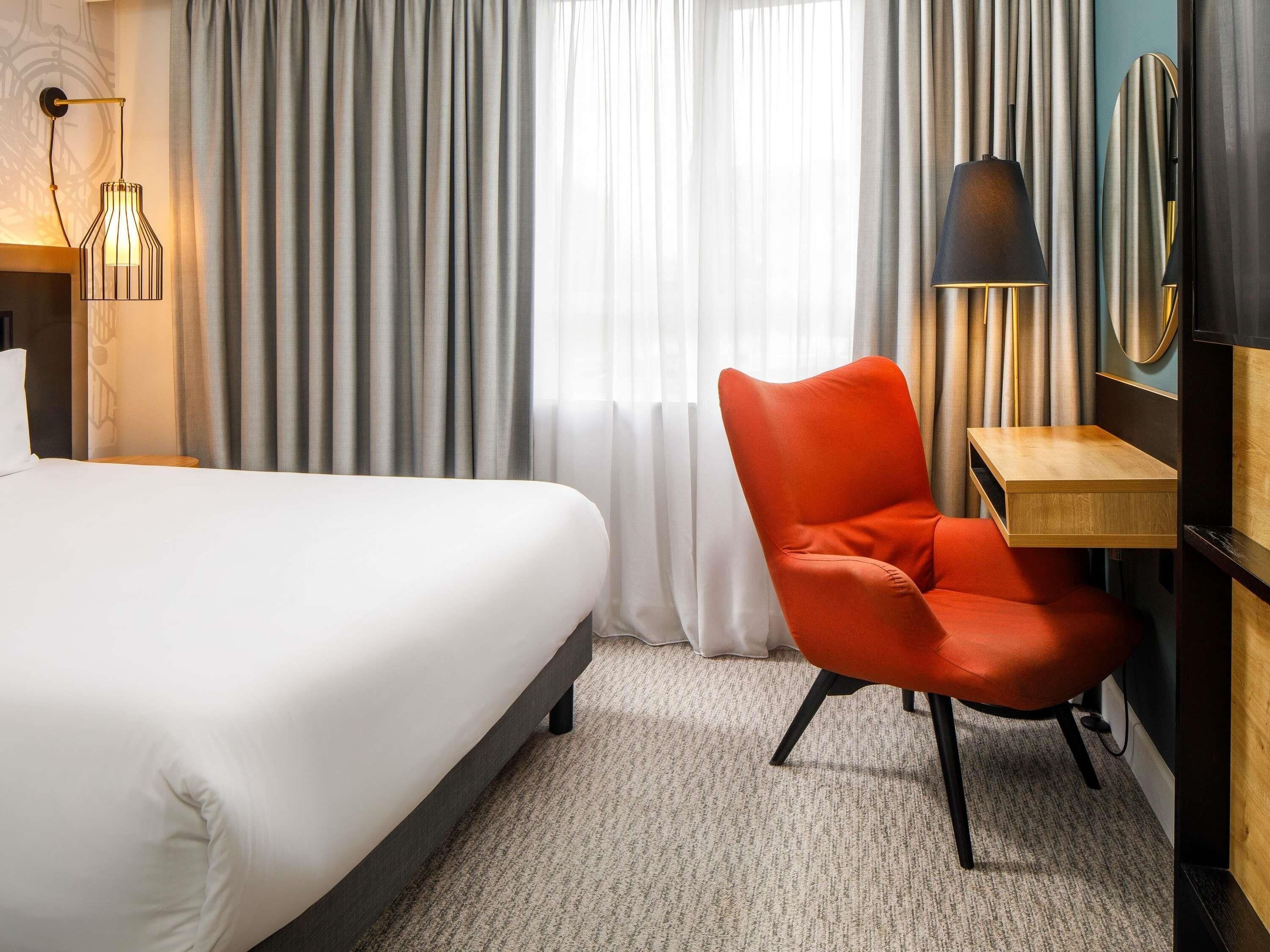 A hotel room with a bed, a red chair, a wooden side table, and a lamp.