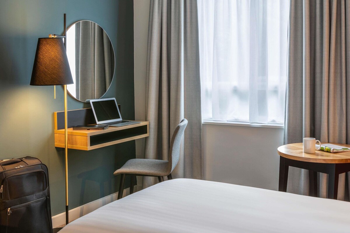Hotelkamer met een bed, een bureau met een laptop, een stoel, een lamp, een koffer en een raam met gordijnen.