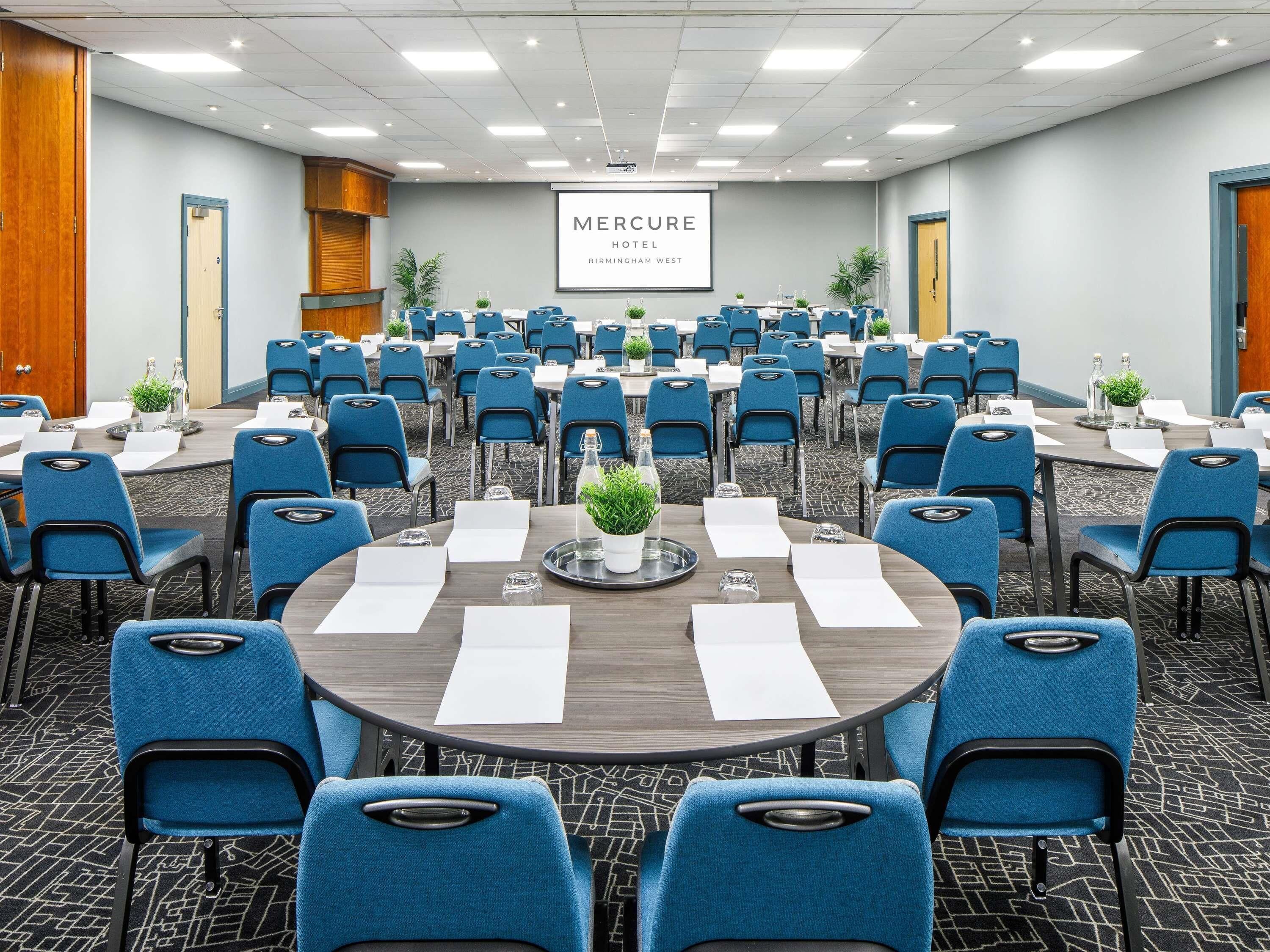 A conference room with round tables, blue chairs, and a large screen displaying 'Mercure Hotel Birmingham West'.