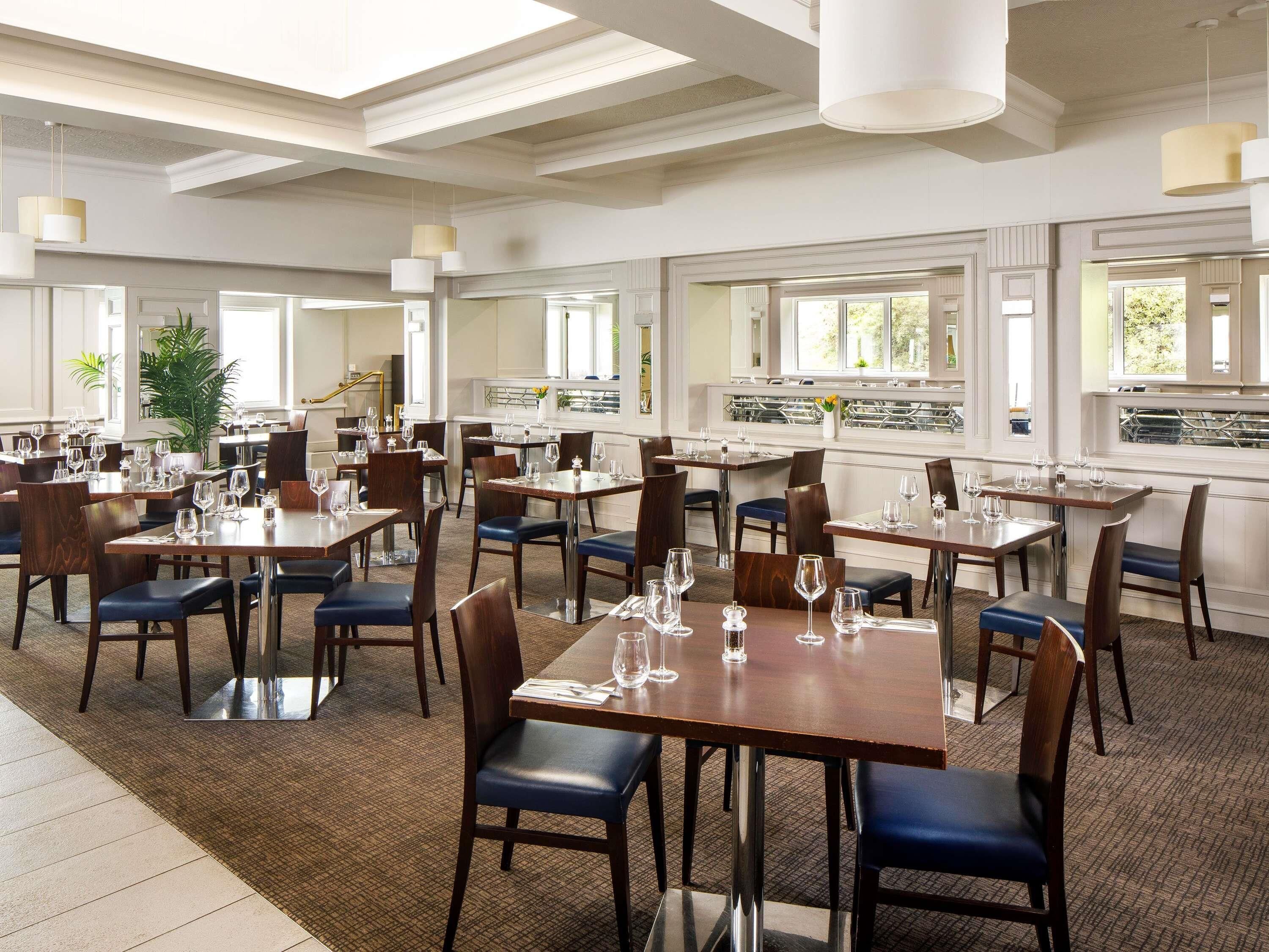 A spacious dining area with multiple tables and chairs, large windows providing natural light, and a modern chandelier.