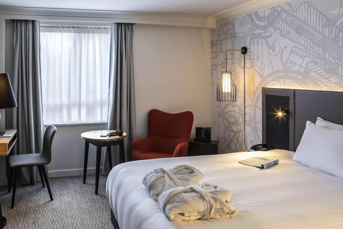 Habitación de hotel con cama, escritorio, silla, mesita, sillón rojo, lámpara y una pared con diseño mecánico.