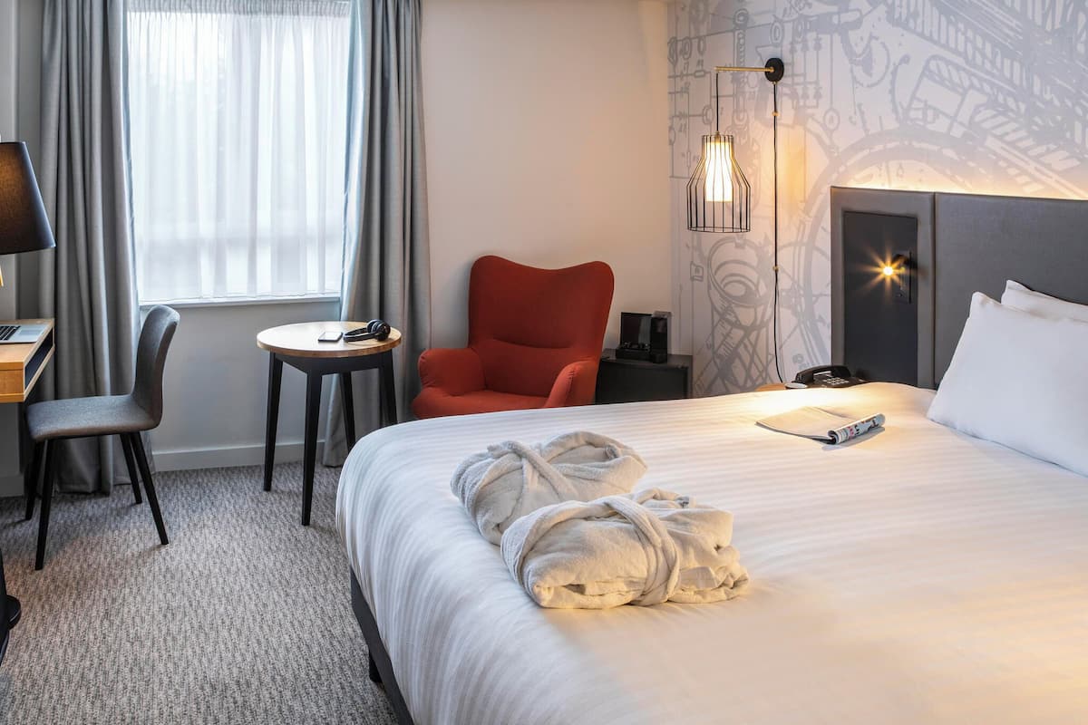 Habitación de hotel con una cama, un escritorio con una computadora portátil, una silla, una mesita y un sillón rojo.