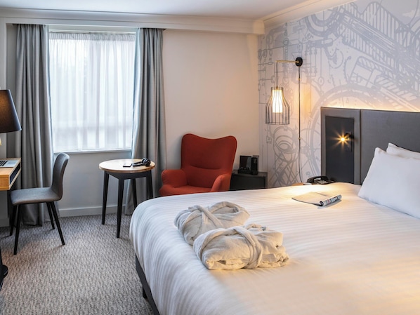 Habitación de hotel con una cama, un escritorio con una computadora portátil, una silla, una mesita y un sillón rojo.