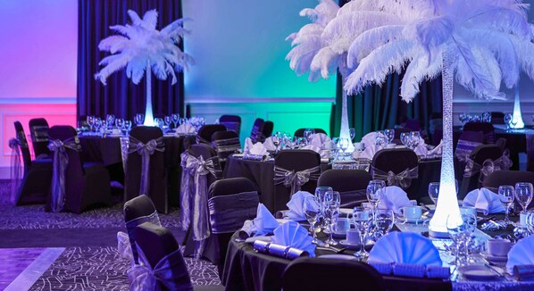 Un comedor formal con mesas dispuestas para una comida, sillas adornadas con lazos y un centro de mesa con un árbol decorativo de plumas.