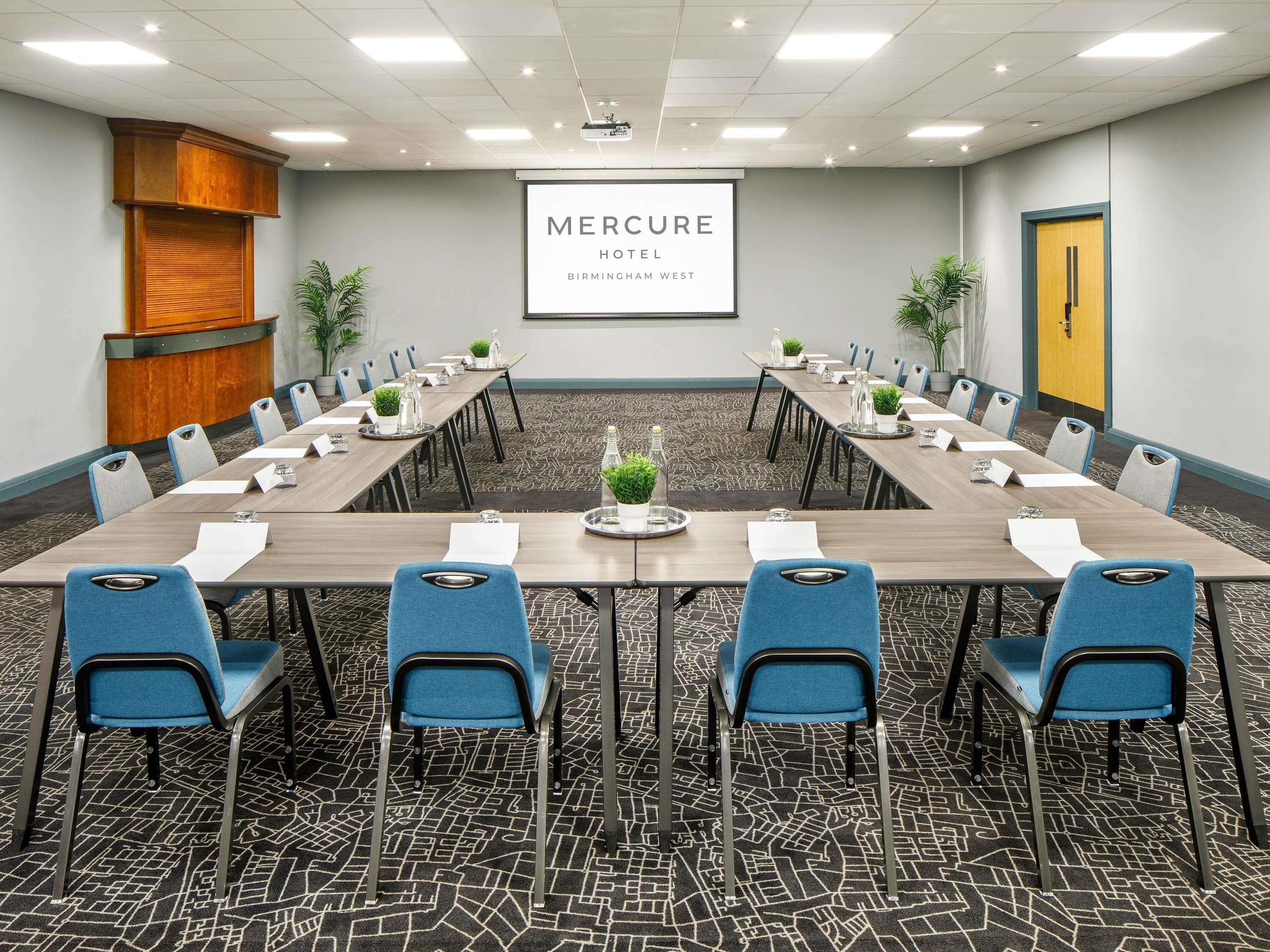 A conference room with a long table, blue chairs, and a projector screen displaying 'Mercure Hotel Birmingham West'.