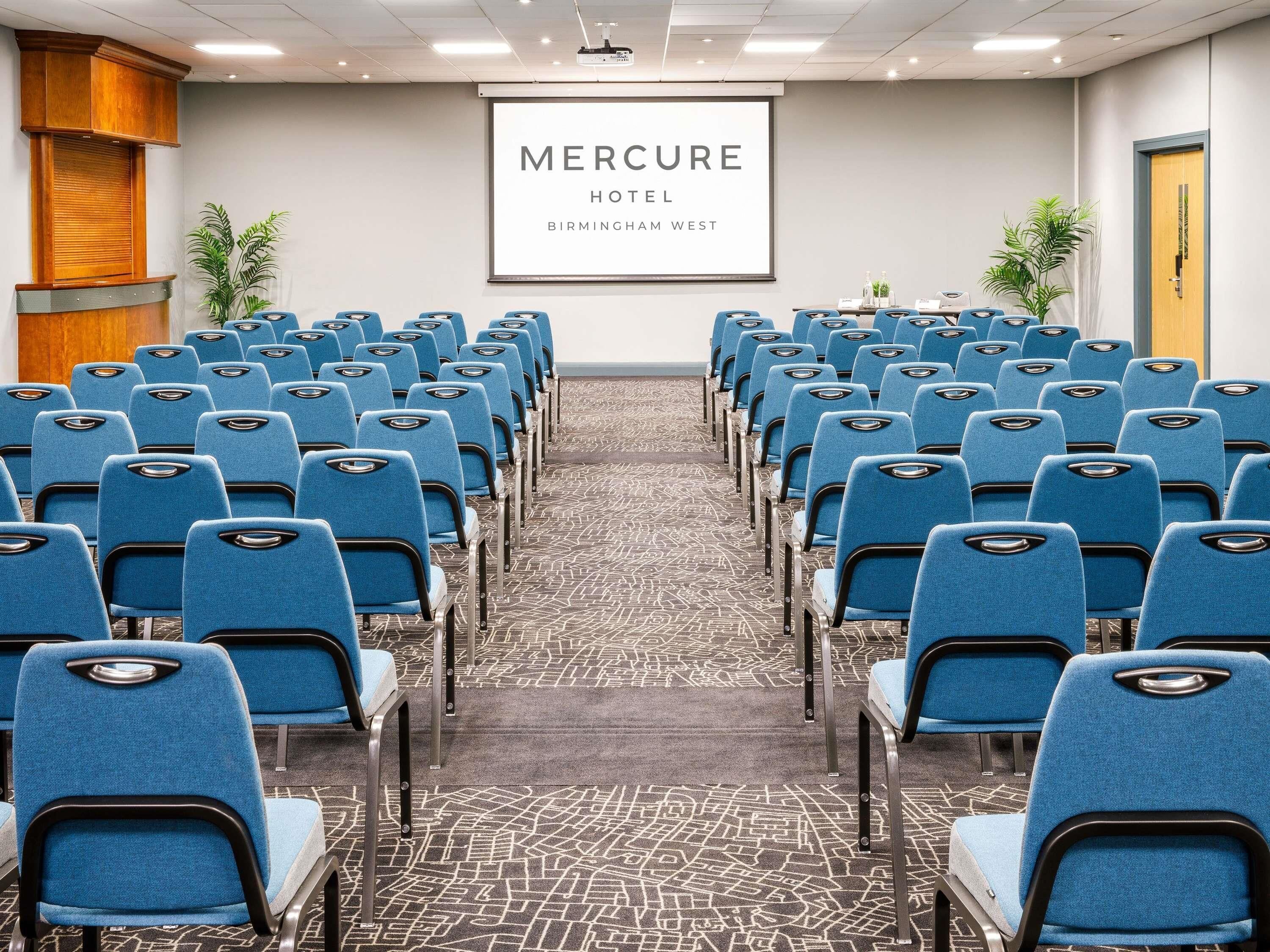 A conference room with rows of blue chairs, a projector screen displaying 'Mercure Hotel Birmingham West', and a carpeted floor.