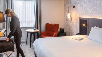 A hotel room with a bed, a desk with a lamp, a chair, a small table, and a wall with a decorative pattern.