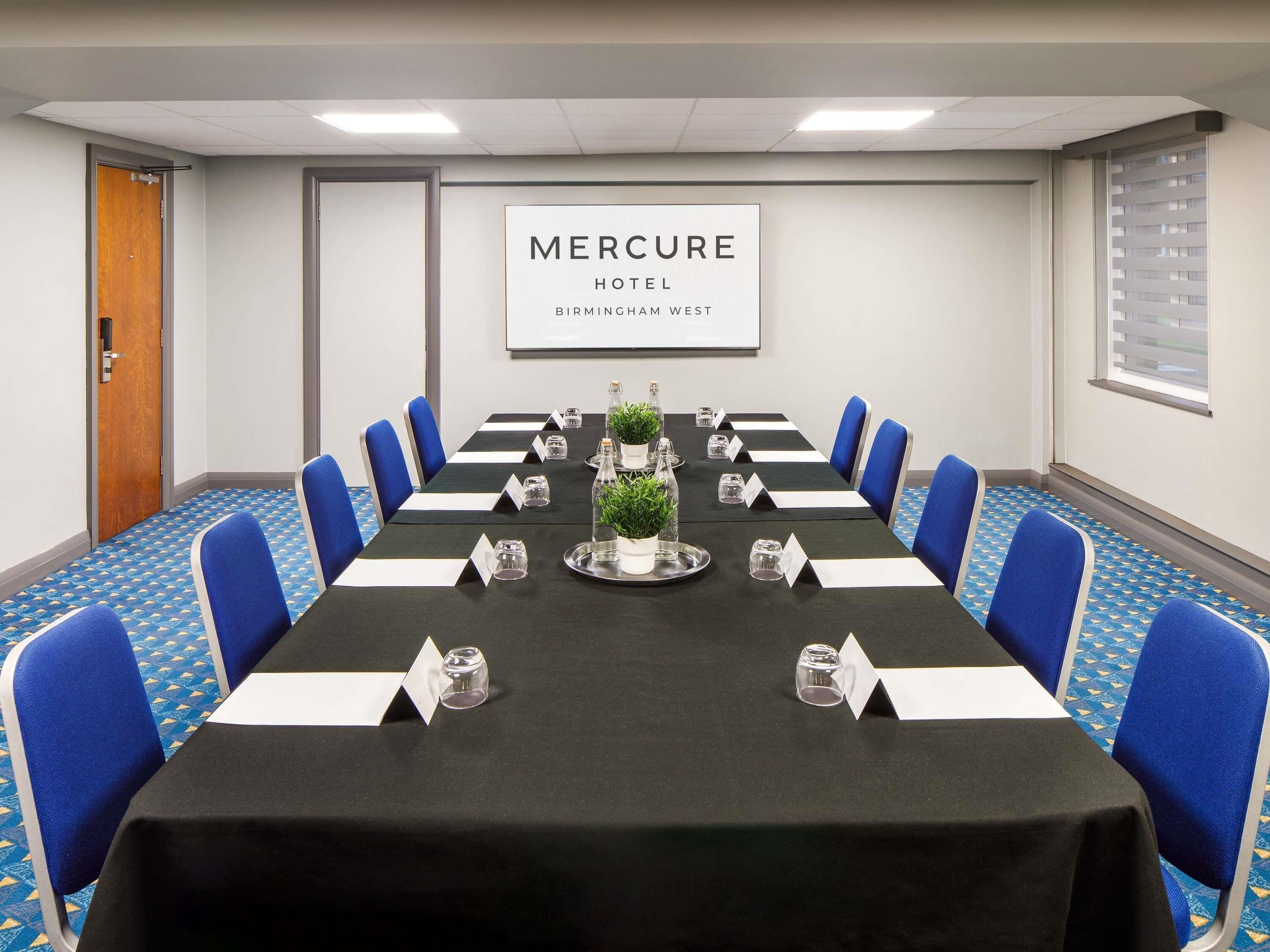 A conference room at Mercure Hotel Birmingham West with a long table, blue chairs, nameplates, water glasses, and a potted plant.
