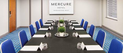 A conference room at Mercure Hotel Birmingham West with a long table, blue chairs, nameplates, water glasses, and a potted plant.