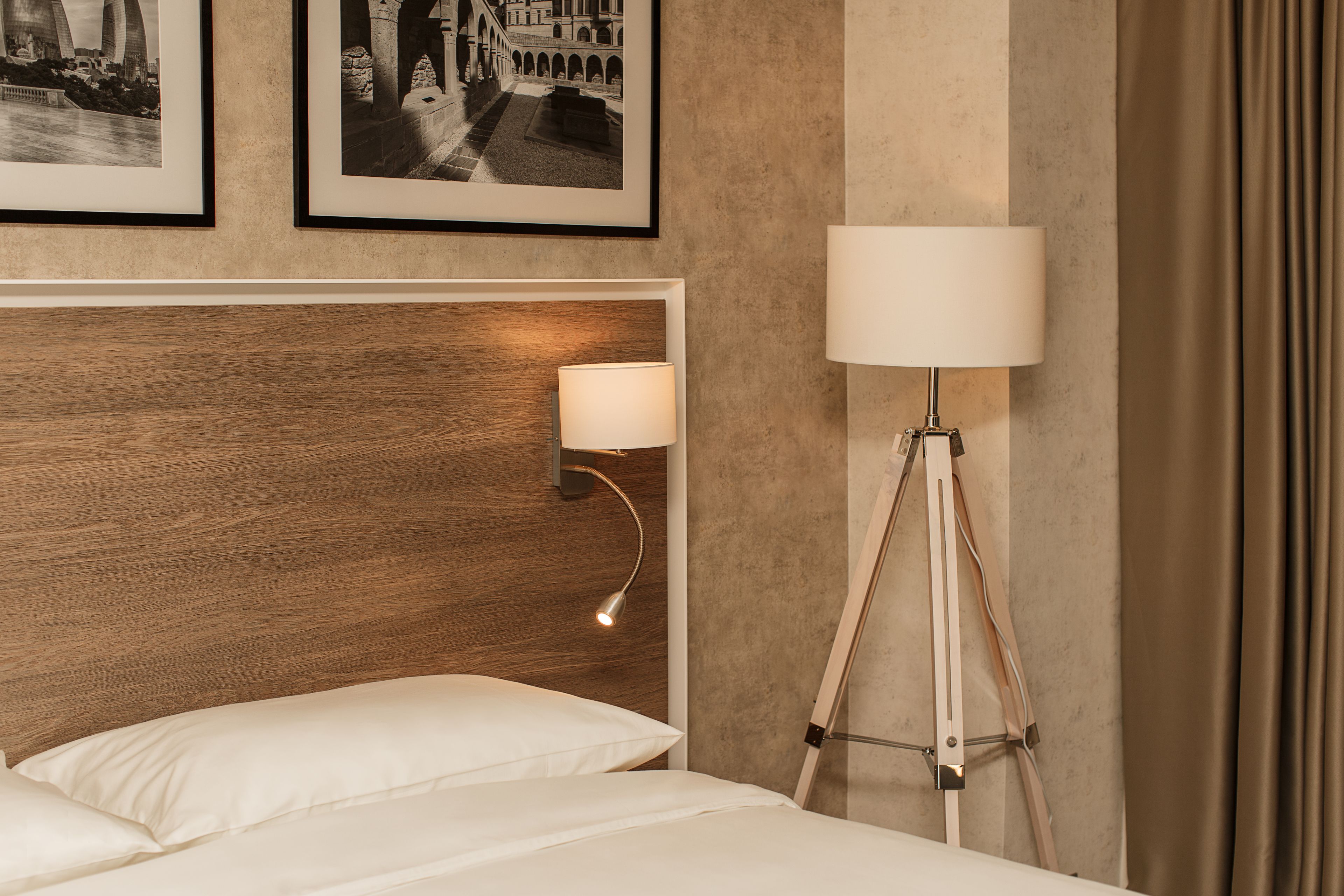 Een hotelkamer met een houten hoofdeinde, een bedlamp, een vloerlamp en twee ingelijste foto’s aan de muur.