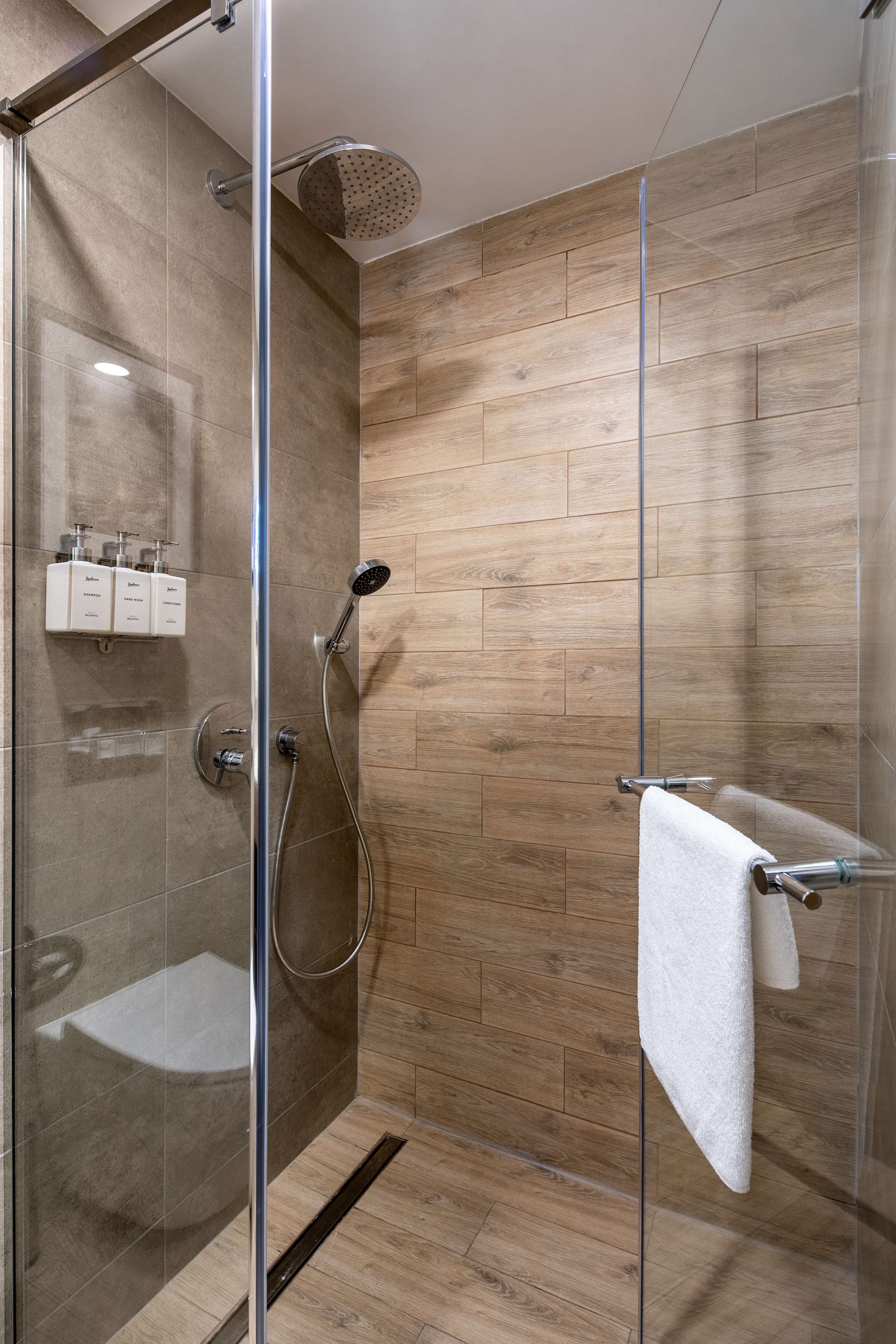 A modern shower with a glass door, wooden wall tiles, and a white towel hanging on a metal towel bar.