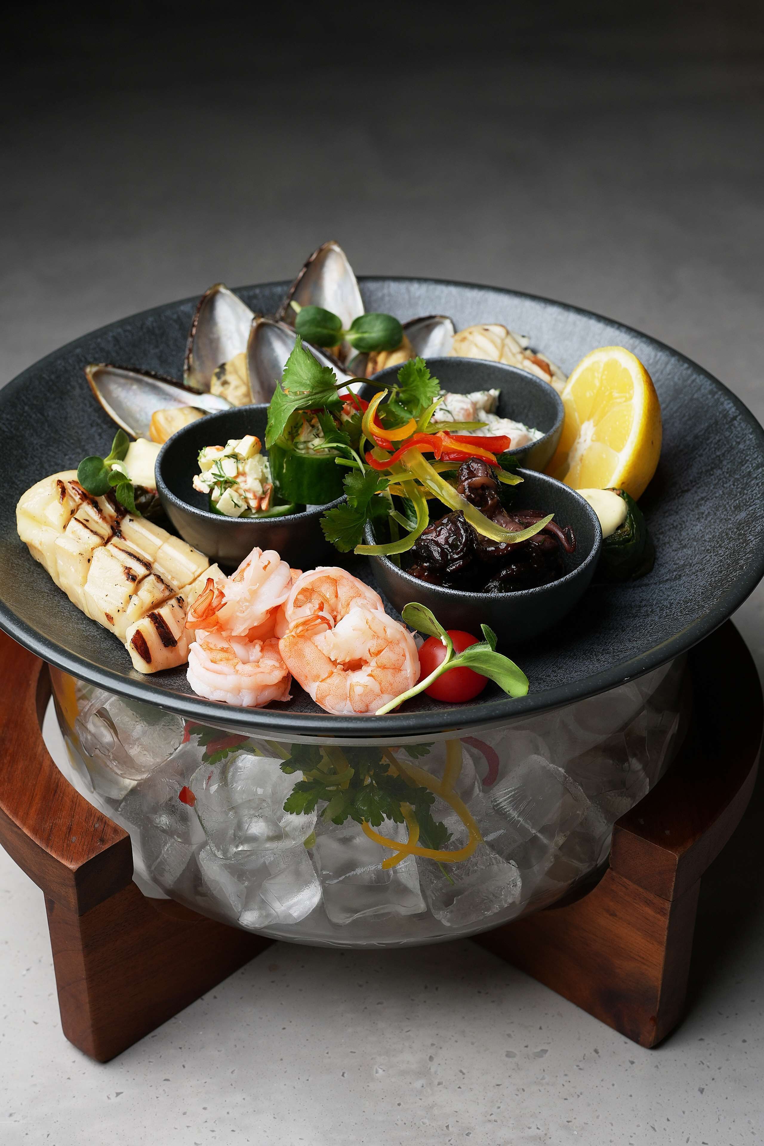 A seafood platter on a dark plate with ice, garnished with herbs and lemon slices.