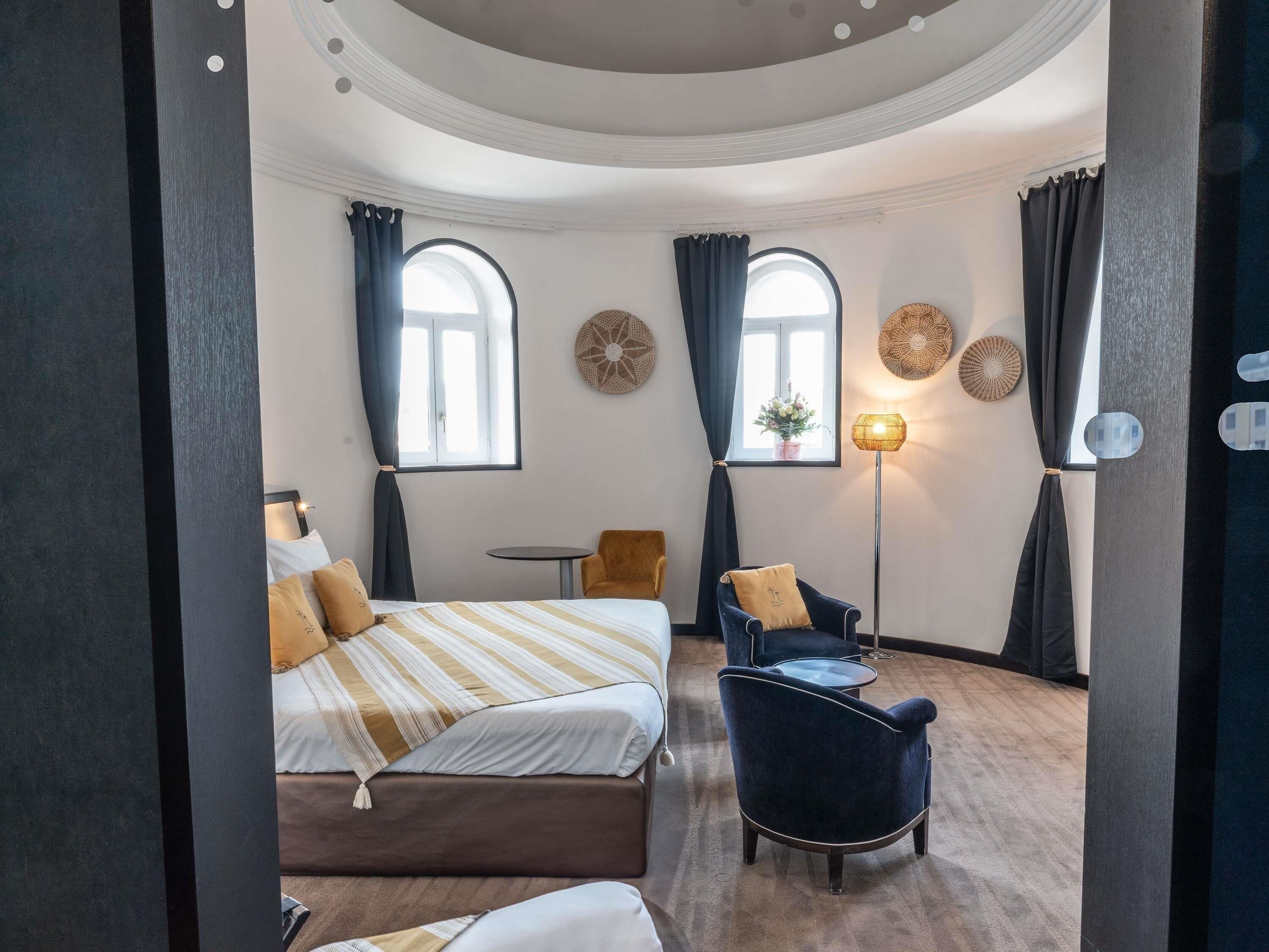 Una habitación de hotel con una cama, una silla, una mesita y una lámpara. Hay dos ventanas con cortinas, y una decoración en la pared sobre las ventanas.