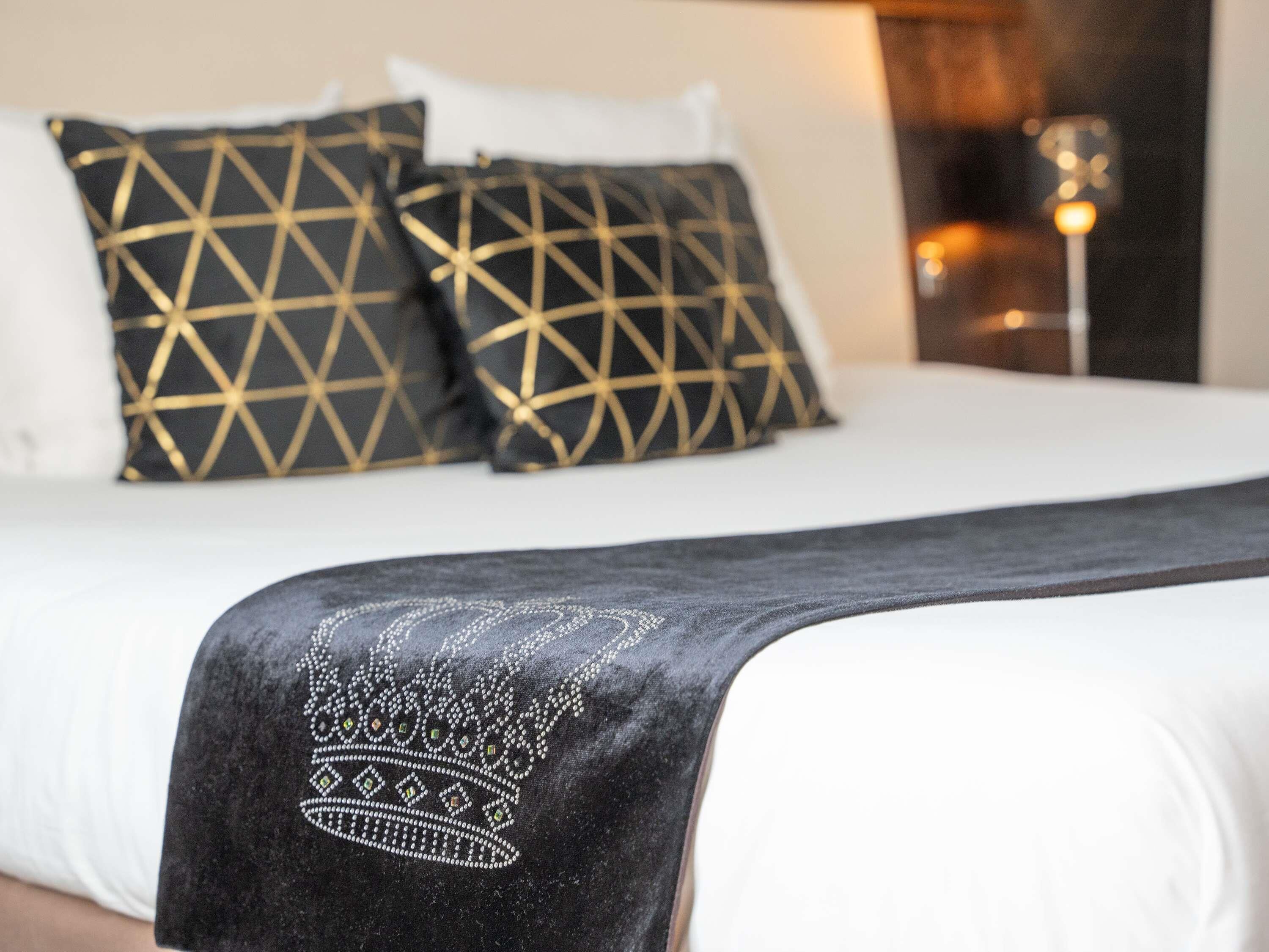Een bed met een kussen met een zwart en gouden patroon, een wit kussen, en een zwart sprei met een kroonmotief.