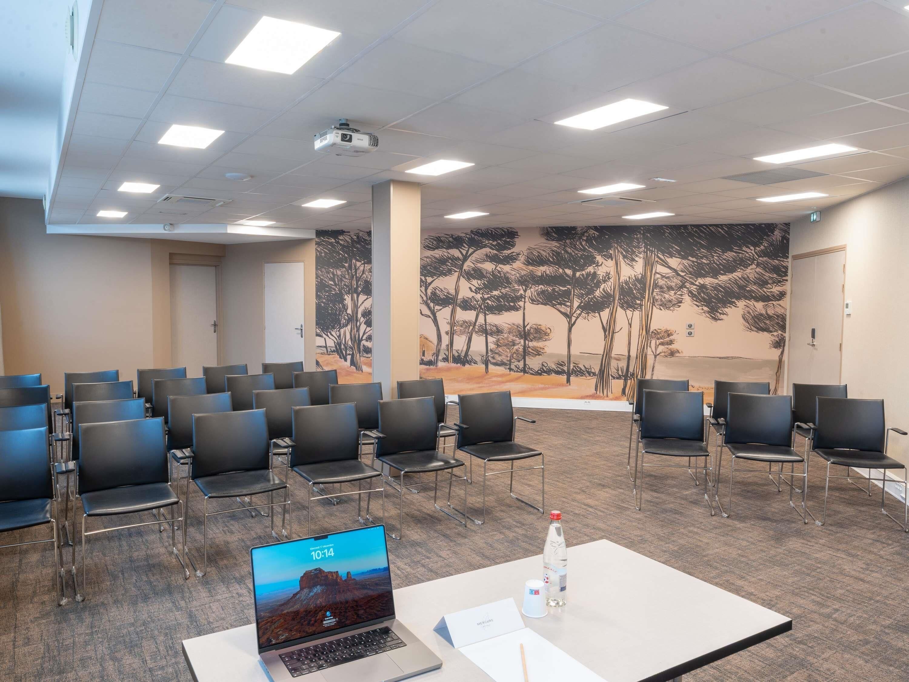 Une salle de conférence avec des rangées de chaises, un projecteur, un ordinateur portable et une bouteille d’eau posée sur une table.
