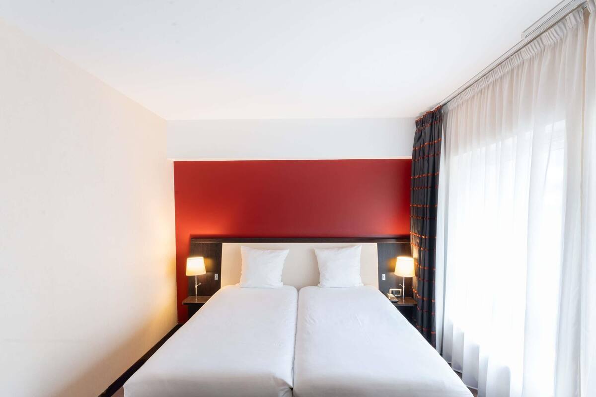 A double bed with a red headboard, two bedside lamps, and sheer curtains.