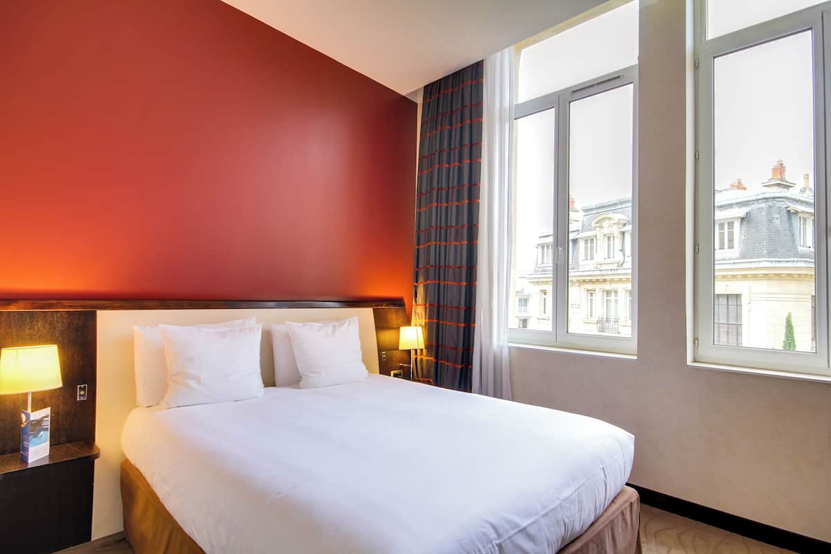 A hotel room with a large bed, two bedside tables with lamps, a window with a view of buildings, and a red accent wall.