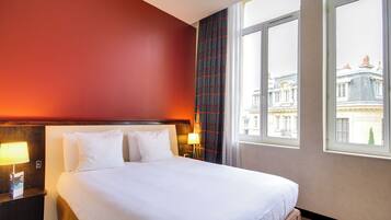 Une chambre d’hôtel avec un grand lit, deux tables de chevet avec des lampes, une fenêtre donnant sur des immeubles et un mur d’accent de couleur rouge.