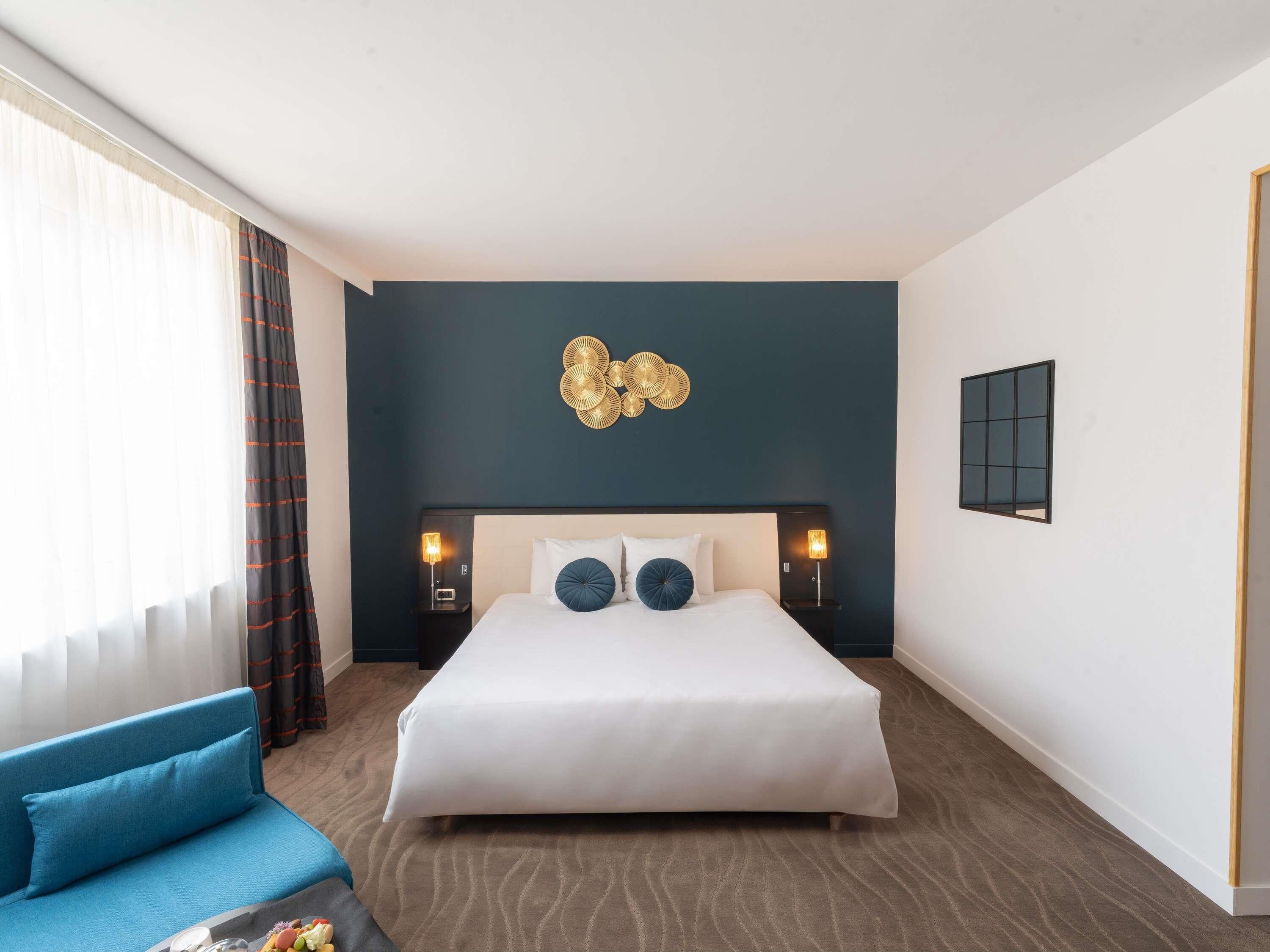 A hotel room with a large bed, a blue sofa, a decorative wall piece, and a window with curtains.