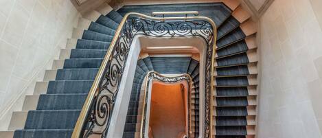 Una imponente escalera con barandales ornamentales en negro y dorado, un rellano central abierto y un vitral en la parte superior.