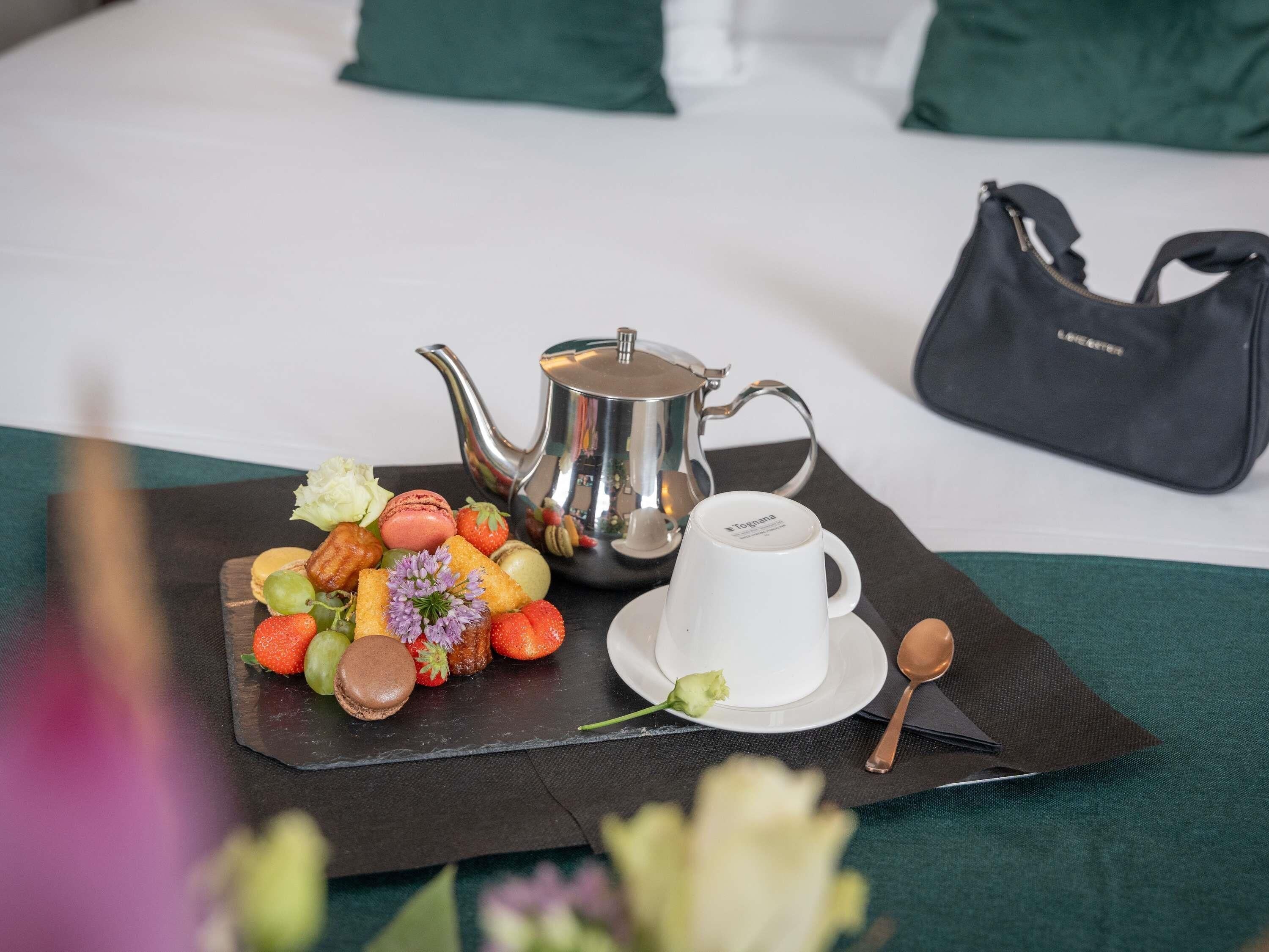 A tray with food, a teapot, and a cup on a table with a bed and a handbag in the background.