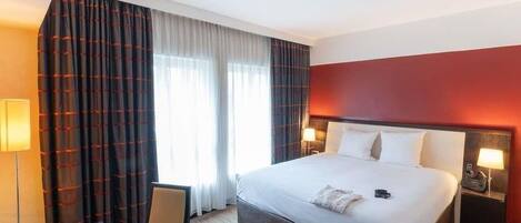 A hotel room with a bed, a desk with a laptop, a chair, a nightstand with a lamp, and a window with curtains.