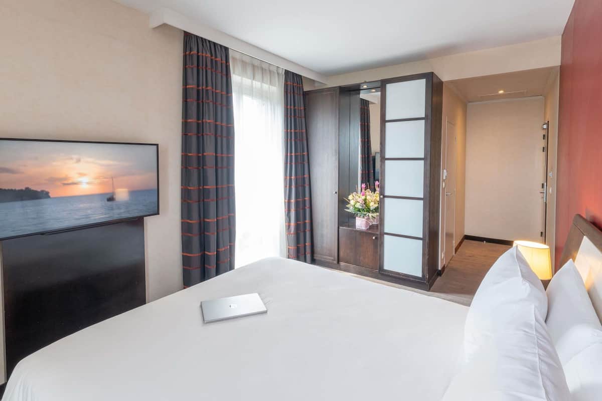 A hotel room with a bed, a laptop, a TV displaying a sunset, and a view of the sea.
