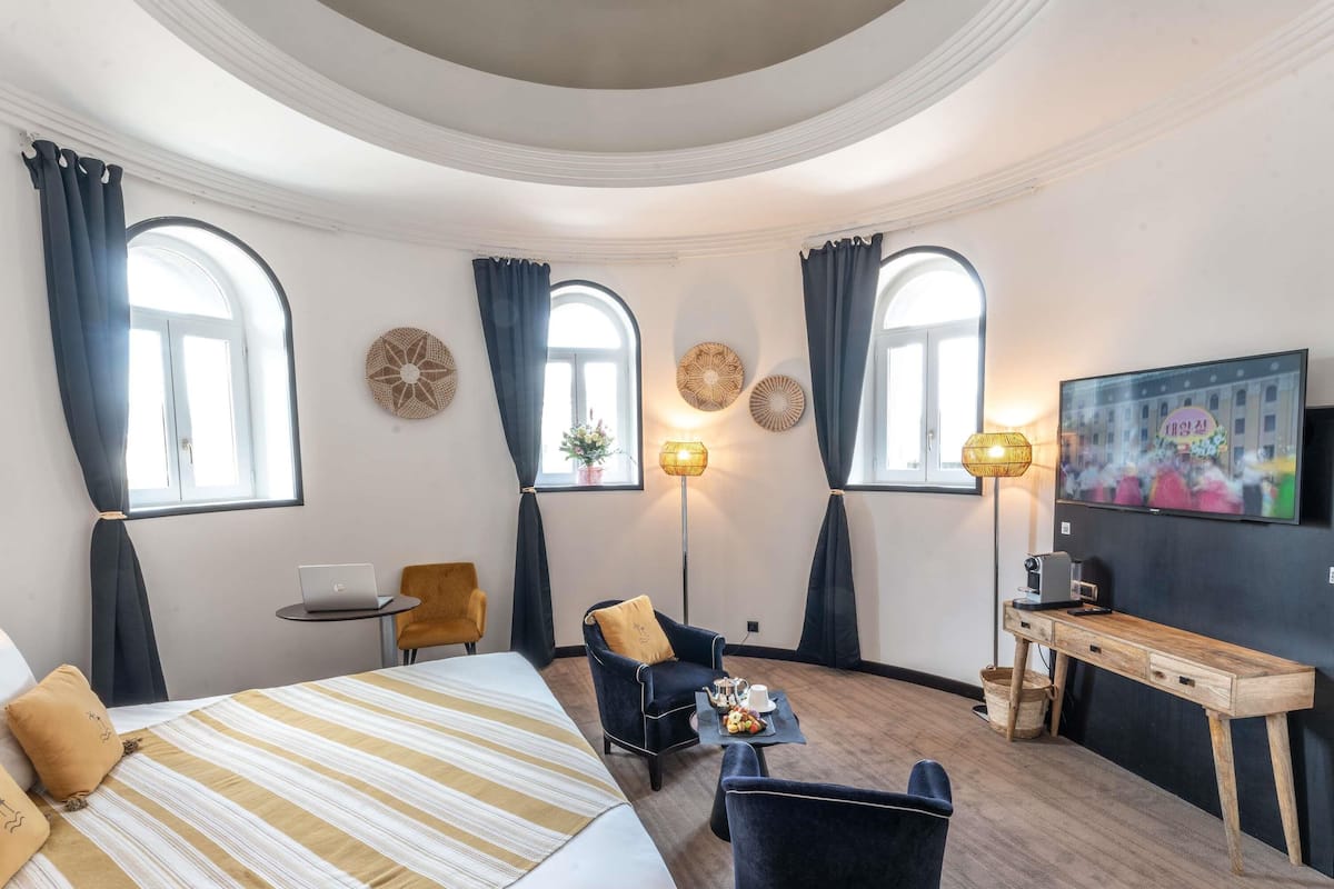 A hotel room with a bed, a desk, a chair, a TV, and two windows with curtains.