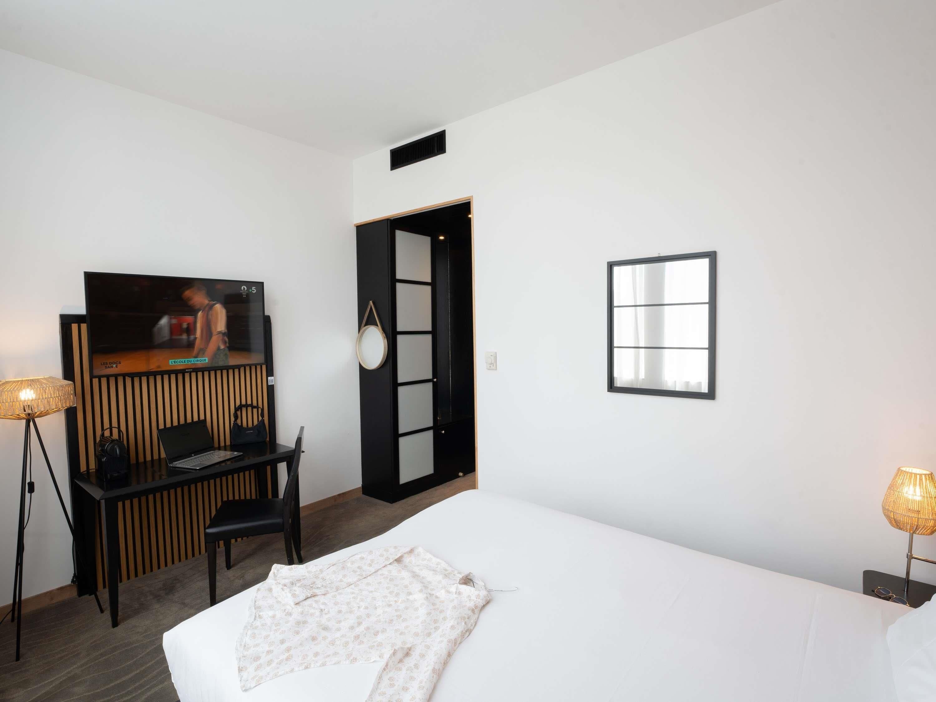 Une chambre d’hôtel avec un lit, un bureau avec un ordinateur portable, une chaise, une télévision et une fenêtre.