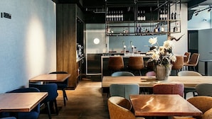 A restaurant with wooden tables and chairs, a bar area with shelves of bottles, and a decorative vase with flowers. - Amsterdam Beach Hotel (Zandvoort)
