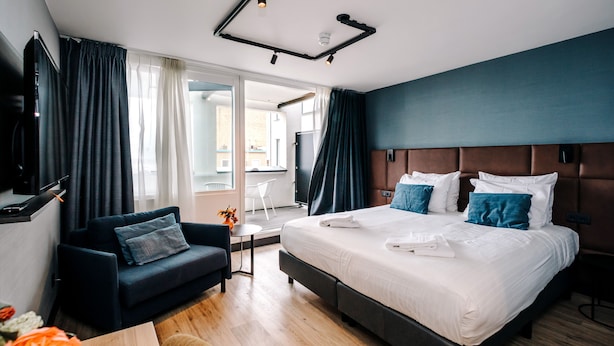 A modern hotel room with a large bed, dark blue sofa, and a small table with a vase of flowers. - Amsterdam Beach Hotel (Zandvoort)