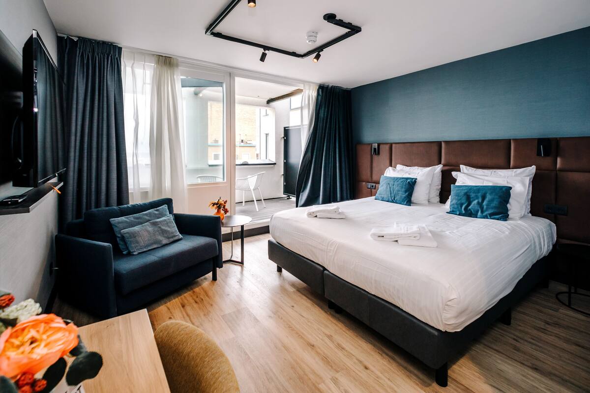 Une chambre d’hôtel moderne avec un grand lit, un canapé bleu foncé et une petite table sur laquelle est posé un vase de fleurs.