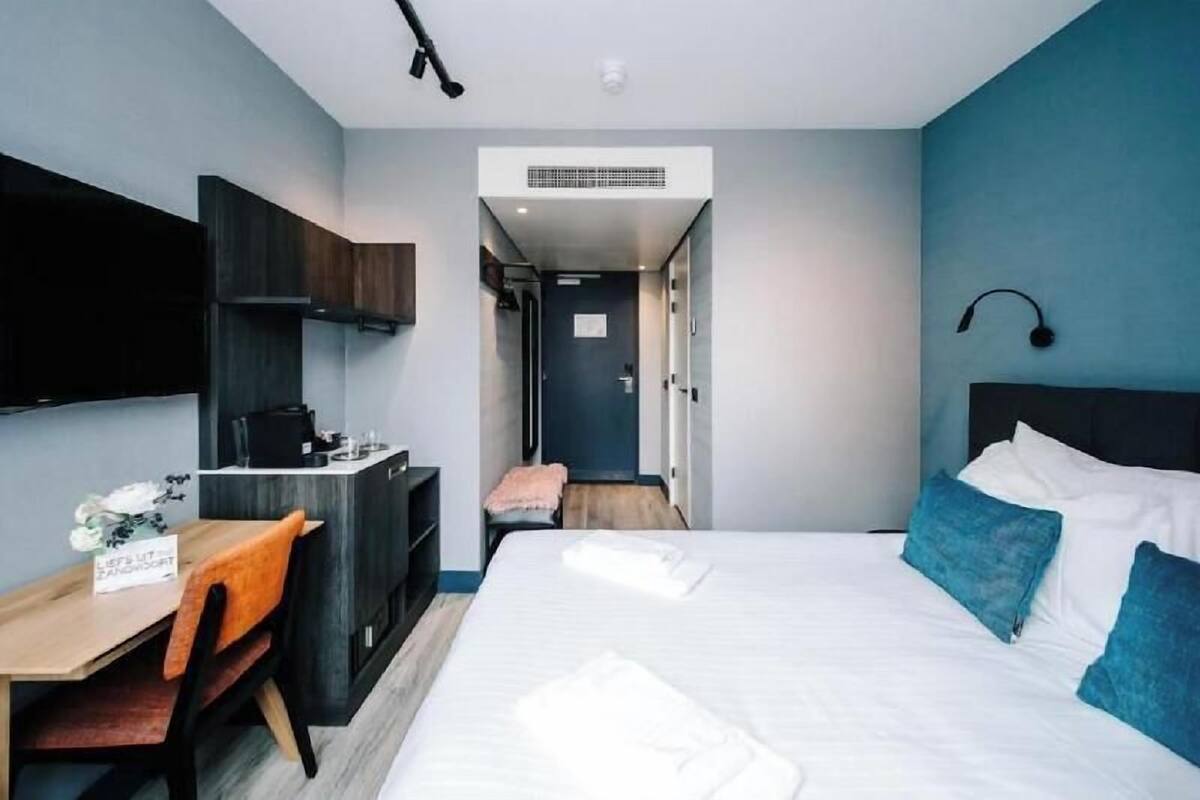 A hotel room with a bed, a desk with a chair, a kitchenette, and a bathroom visible through an open door.