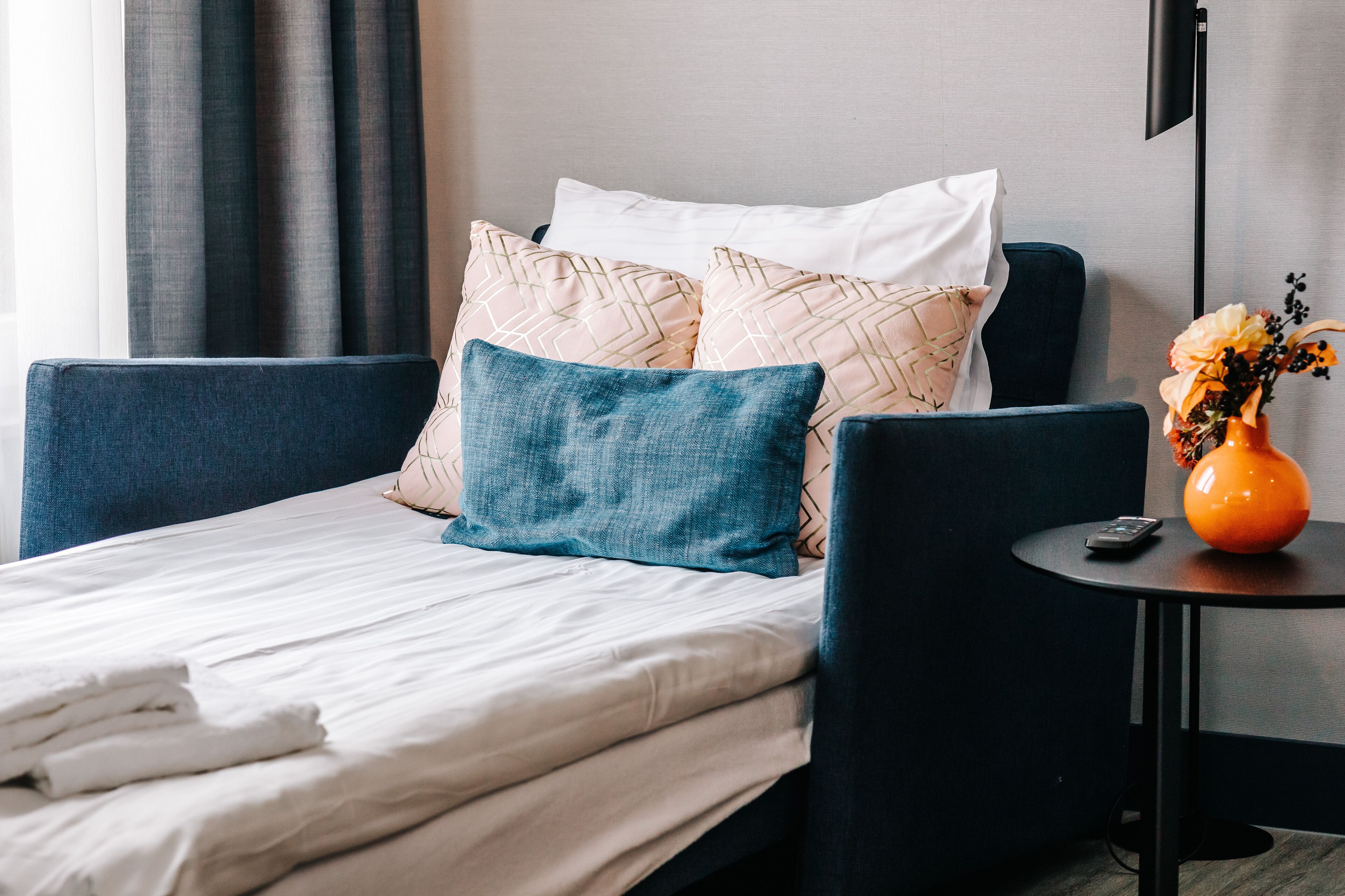 A neatly made bed with white linens, a blue upholstered headboard, and decorative pillows. A small round table with a vase of flowers and a remote control is beside the bed.