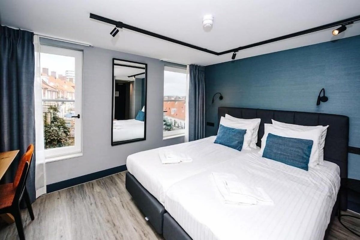 A hotel room with a bed, a desk, a chair, a window with curtains, and a view of buildings outside.