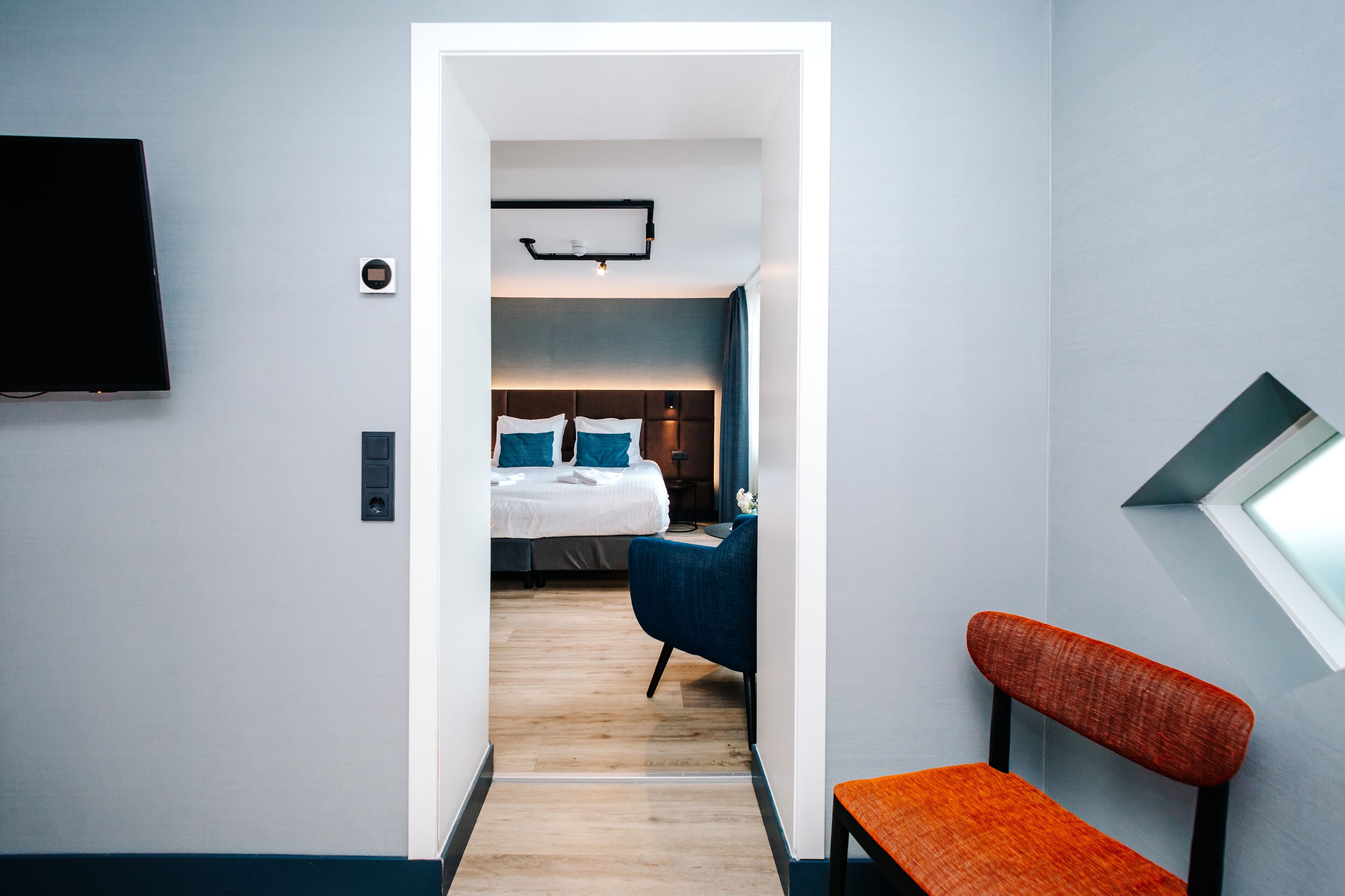 Ein modernes Hotelzimmer mit Holzboden, einem Bett mit blau-weißer Bettwäsche, einem blauen Sessel und einem kleinen Tisch.