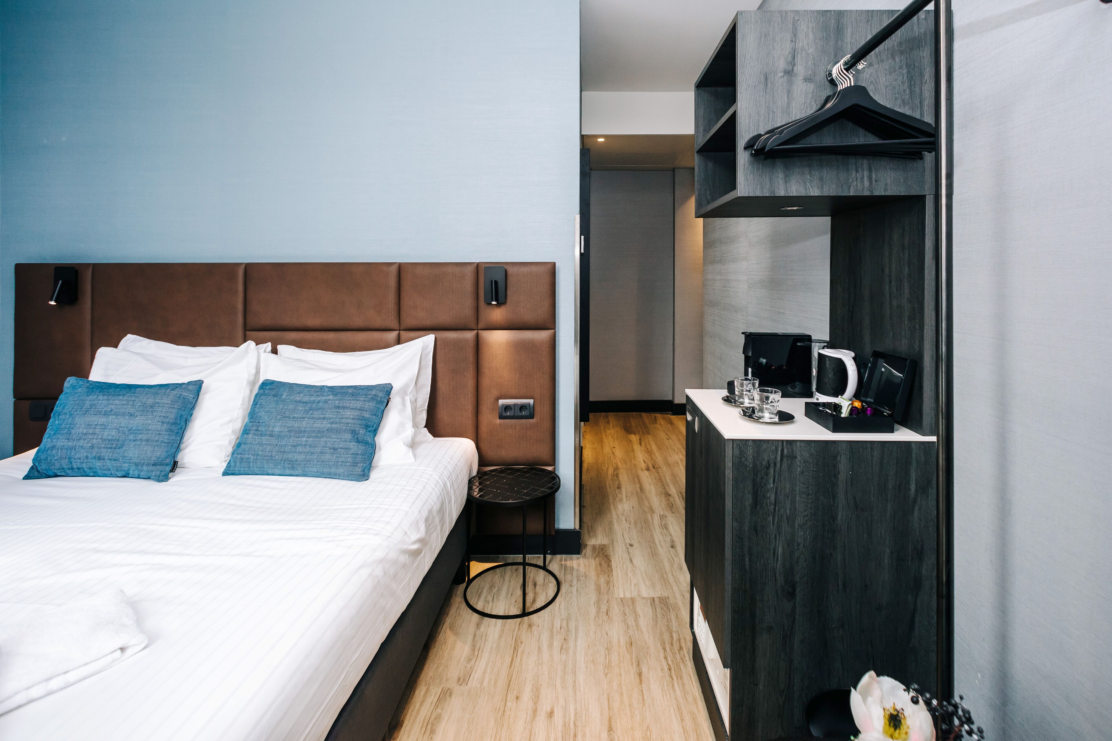 A modern hotel room with a large bed, a small bedside table, a coffee machine, and a wardrobe with a mirror.