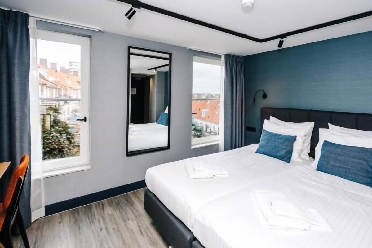A hotel room with a large bed, a desk, a chair, a mirror, and a view of the city.