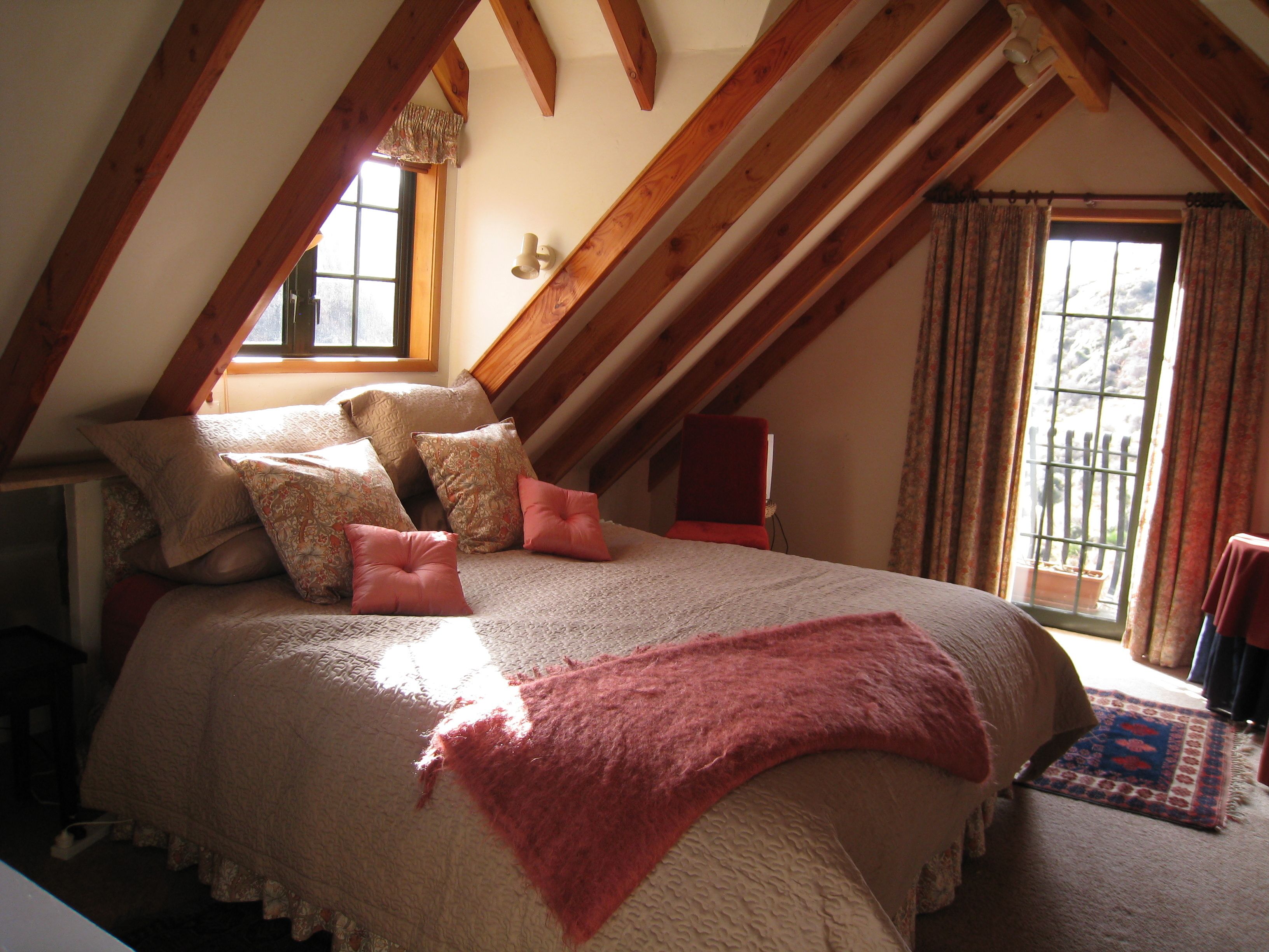 A bedroom with a sloped ceiling, a large bed with multiple pillows, a red chair, and a window with curtains.