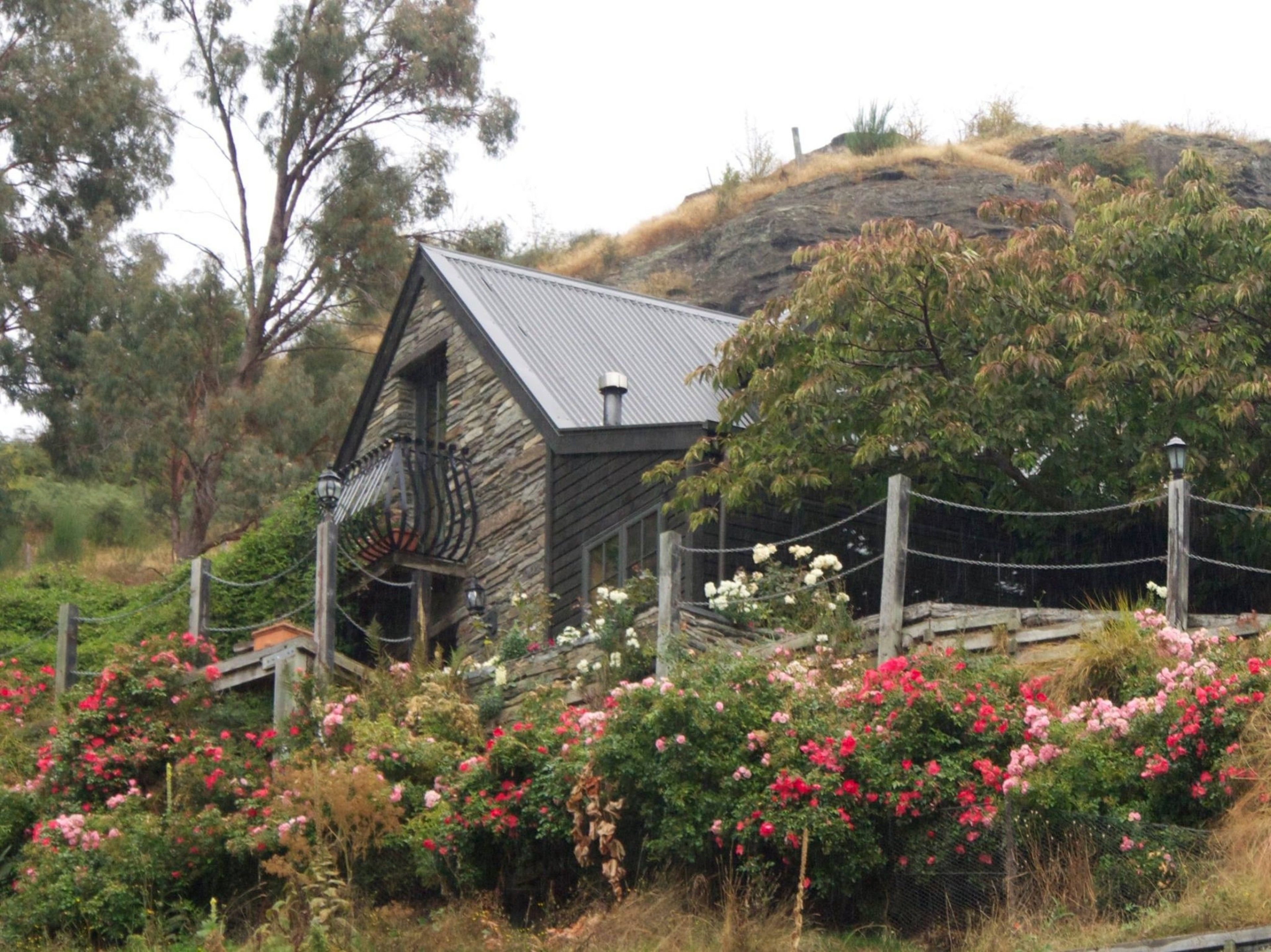 A rustic stone building with a sloped roof, surrounded by a variety of flowering plants and a chain-link fence.