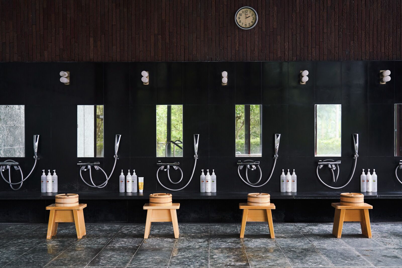 A modern bathroom with multiple shower stations, wooden stools, and a clock on the wall.